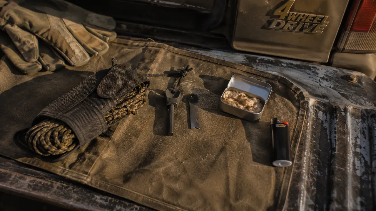 Flat lay of a vehicle fire kit on a tailgate: ferro rod on a short lanyard, striker, small tin of tinder, and a butane lighter