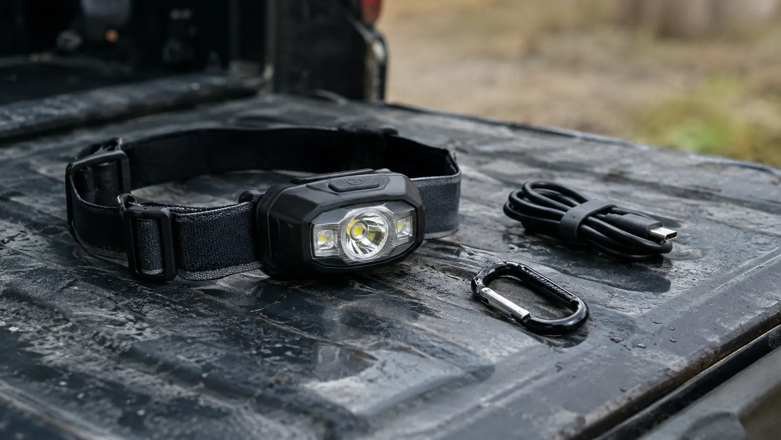 A close view of a headlamp on a metal tailgate next to a coiled USB-C cable and a carabiner, overcast daylight outdoors